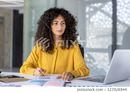 A businesswoman, dressed in yellow, focuses on paperwork with a pen at her desk. A laptop sits nearby, possibly for research or analysis. A businesswoman, dressed in yellow, focuses on paperwork with a pen at her desk. A laptop sits nearby, possibly for research or analysis. 127826944