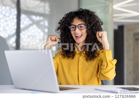 A happy businesswoman celebrates success while looking at her laptop screen in an office setting. She wears glasses and a yellow shirt. 127826966
