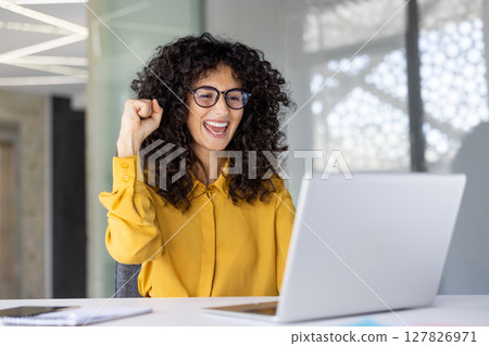 Successful joyful woman celebrating victory and successful achievement results. Businesswoman at workplace working with laptop holding hand up, gesture of triumph. 127826971