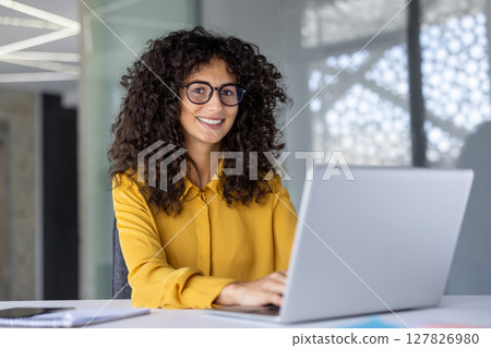 Smiling hispanic businesswoman with curly hair and eyeglasses typing on laptop in office. Confident professional engaged in work, with phone on desk. Represents modern workplace and successful career Smiling hispanic businesswoman with curly hair and eyeglasses typing on laptop in office. Confident professional engaged in work, with phone on desk. Represents modern workplace and successful career 127826980