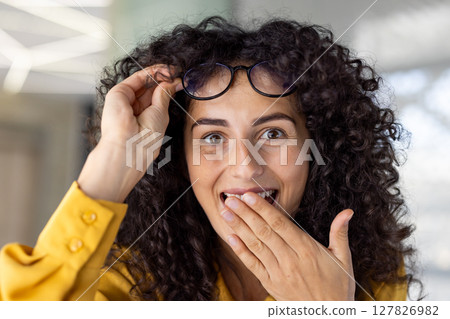 A beautiful woman with curly hair and a yellow blouse, looking surprised as she holds up her glasses indoors, in a close-up portrait 127826982