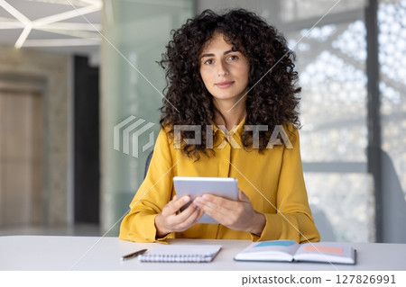 A woman with curly hair sits at a desk in a bright office setting, holding a tablet. She is wearing a yellow shirt and looking at the camera. A woman with curly hair sits at a desk in a bright office setting, holding a tablet. She is wearing a yellow shirt and looking at the camera. 127826991
