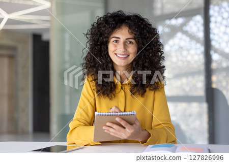 Young woman smiles while taking notes, ready for a virtual meeting at her well-lit workspace 127826996