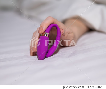 Woman holding curved purple vibrator while lying in bed. Woman holding curved purple vibrator while lying in bed. 127827020