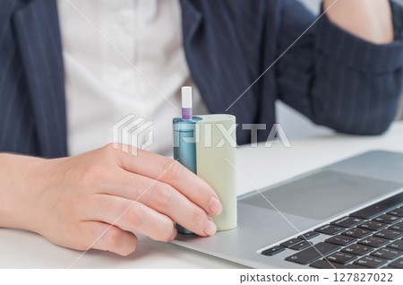Business woman smoking an electronic cigarette while working on a laptop. Tobacco heating system. Business woman smoking an electronic cigarette while working on a laptop. Tobacco heating system. 127827022