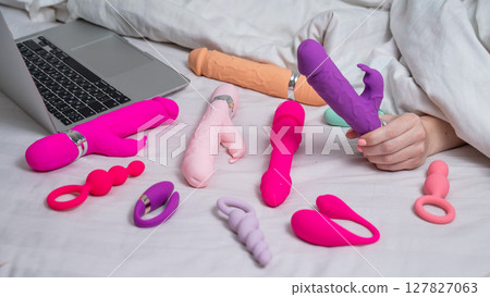 Woman holding sex toy. Various vibrators and laptop on bed.  127827063