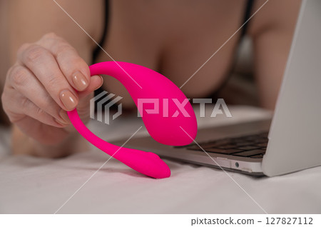 A woman is lying in bed holding a curved vibrator and looking at a laptop.  127827112