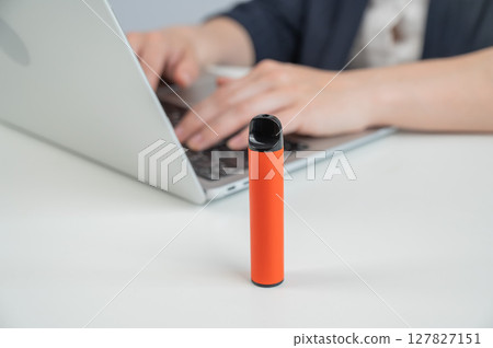 Caucasian businesswoman working on laptop. Disposable vape in foreground. Caucasian businesswoman working on laptop. Disposable vape in foreground. 127827151