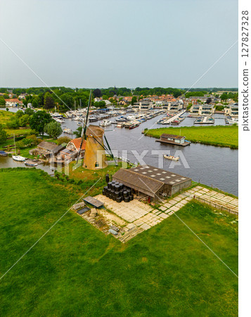 A top-down view of a marina with multiple boats docked along wooden piers, a boat cruising through a narrow waterway surrounded by green parks and natural grassy areas. Windmills. A top-down view of a marina with multiple boats docked along wooden piers, a boat cruising through a narrow waterway surrounded by green parks and natural grassy areas. Windmills. 127827328