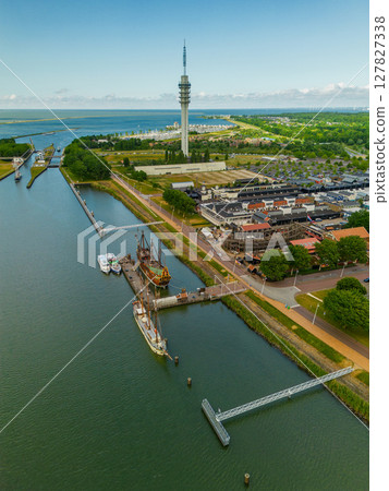 An aerial view of a coastal town with a harbor, colorful buildings, a tall communication tower, and a replica ship docked along the water. Surrounding areas include parking lots and green spaces. An aerial view of a coastal town with a harbor, colorful buildings, a tall communication tower, and a replica ship docked along the water. Surrounding areas include parking lots and green spaces. 127827338