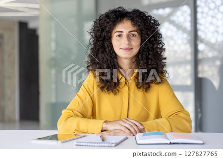A smiling woman with curly hair sits at a desk with a tablet, notebook and pen in front of her in a bright, modern office space 127827765