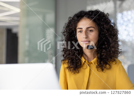 A woman with curly hair wearing a headset, looking attentively at a computer screen, likely providing customer service or support in a modern office. 127827860