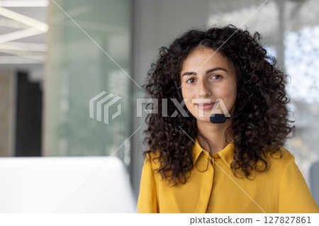 A smiling woman with curly hair wears a headset. She is likely working in a customer service or call center environment. A smiling woman with curly hair wears a headset. She is likely working in a customer service or call center environment. 127827861