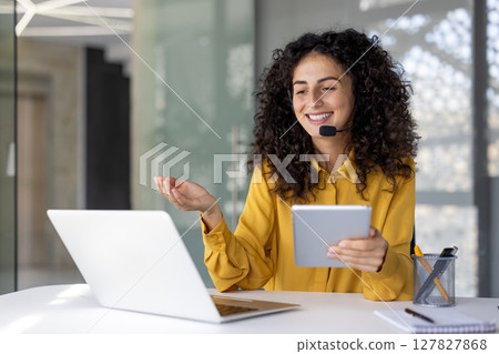 Support service worker, businesswoman with headset and tablet in hands communicates and consults clients remotely, office worker smiles satisfied uses laptop inside office. Support service worker, businesswoman with headset and tablet in hands communicates and consults clients remotely, office worker smiles satisfied uses laptop inside office. 127827868