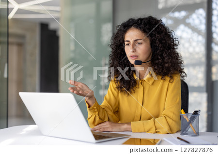 A woman with curly hair wearing a headset and yellow shirt, looks at a laptop while gesturing, indicating a business meeting or customer service interaction. A woman with curly hair wearing a headset and yellow shirt, looks at a laptop while gesturing, indicating a business meeting or customer service interaction. 127827876