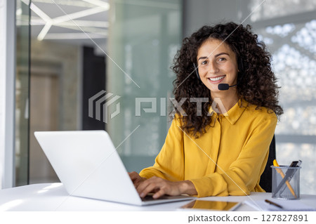 Hispanic businesswoman working on laptop in office environment. Wearing headset, demonstrating teamwork, communication skills. Engaged in customer service, business management, project organization. 127827891