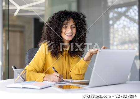 Smiling woman in yellow shirt is attending an online course, taking notes while gesturing in front of her laptop Smiling woman in yellow shirt is attending an online course, taking notes while gesturing in front of her laptop 127827894