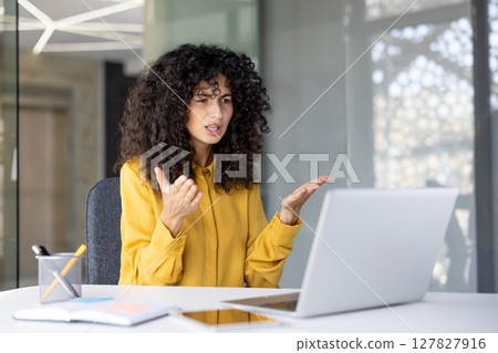 A woman with curly hair appears frustrated during a video call, gesturing with her hands while seated in front of a laptop. 127827916