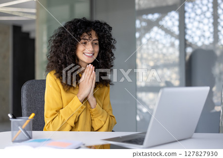 A happy woman in a yellow shirt smiles while looking at her laptop with her hands together, as if in prayer. A happy woman in a yellow shirt smiles while looking at her laptop with her hands together, as if in prayer. 127827930