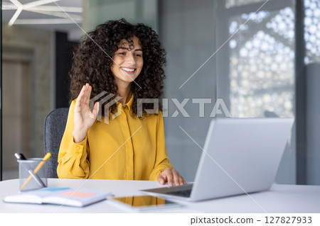 Hispanic businesswoman participating in virtual meeting using laptop in professional office. Woman waves and smiles, conveying friendly communication. Hispanic businesswoman participating in virtual meeting using laptop in professional office. Woman waves and smiles, conveying friendly communication. 127827933
