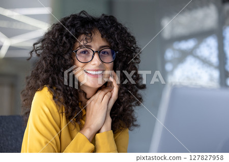 Hispanic businesswoman with curly hair and glasses smiling confidently in office environment. She exudes professionalism and positivity. Ideal image for concepts of success, confidence, and corporate 127827958