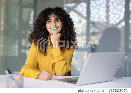A smiling woman with curly hair leans on her hand, sitting at a desk with a laptop, looking towards the camera. 127828071