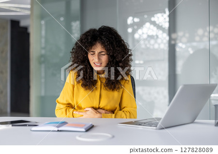Hispanic businesswoman sits at office desk, experiencing stomach pain. She holds her abdomen, showing stress and discomfort. Nearby are laptop and notebook, indicating a work setting. 127828089