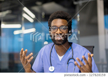 An African-American male doctor wearing glasses and a headset smiles while gesturing during a telehealth consultation. A stethoscope is around his neck. 127828159