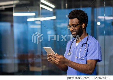 Young successful experienced doctor standing inside the clinic with a tablet computer in his hands. The doctor reviews patients' data and communicates with clients remotely. Young successful experienced doctor standing inside the clinic with a tablet computer in his hands. The doctor reviews patients' data and communicates with clients remotely. 127828189
