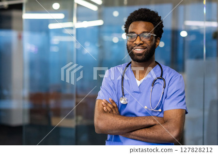 A smiling African American doctor wearing glasses, a stethoscope, and scrubs, standing in a medical setting with arms crossed. A smiling African American doctor wearing glasses, a stethoscope, and scrubs, standing in a medical setting with arms crossed. 127828212