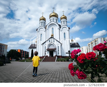 Child in yellow T-shirt is standing in front of Orthodox church in summer. View from back. Golden domes. Church on sunny day. child in front of the church. 127828309