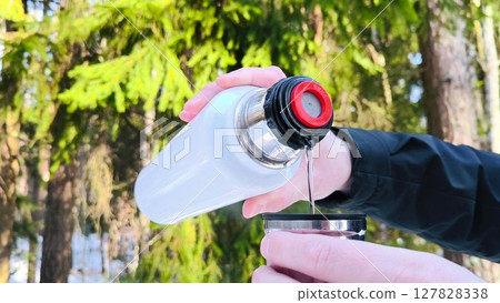 Hot drink flows from thermos in snowy forest in winter. Water flows, pours hot drink against backdrop of of winter nature. Active lifestyle and travel concept. man pours hot drink from thermos 127828338