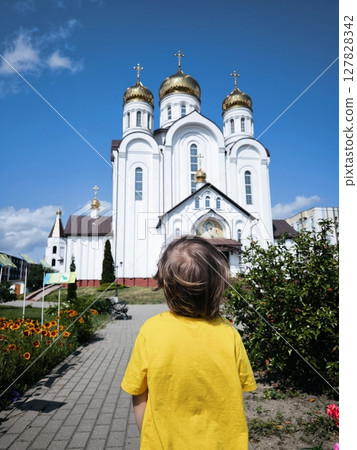 Child in yellow T-shirt is standing in front of Orthodox church in summer. View from back. Golden domes. Church on sunny day. child in front of the church. 127828342