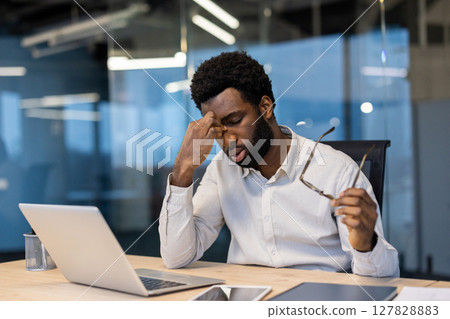 Tired and overworked man sitting at desk, businessman with dizziness sitting with laptop, eye pain, late work until evening time. Tired and overworked man sitting at desk, businessman with dizziness sitting with laptop, eye pain, late work until evening time. 127828883