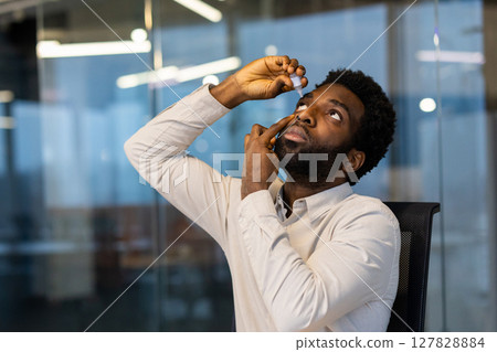 A man applies eye drops while looking up in a modern office setting, highlighting healthcare and wellness. A man applies eye drops while looking up in a modern office setting, highlighting healthcare and wellness. 127828884