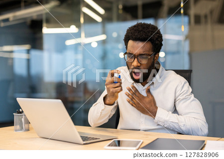 An african american man in an office uses an inhaler, potentially experiencing an asthma attack while working on a laptop. An african american man in an office uses an inhaler, potentially experiencing an asthma attack while working on a laptop. 127828906