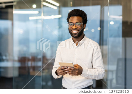 Portrait of a young successful businessman inside the office at his workplace. The man is smiling and looking at the camera, holding a tablet computer in his hands. 127828910