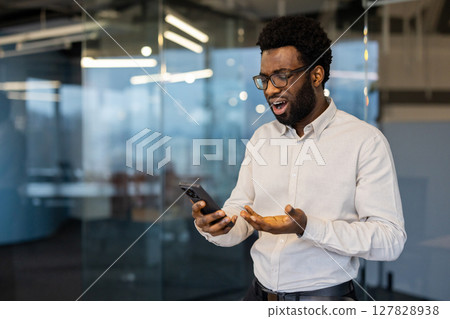 An African American man with glasses looks shocked at something he sees on his phone while in an office setting. An African American man with glasses looks shocked at something he sees on his phone while in an office setting. 127828938