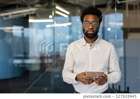 Serious focused man looking at camera. Businessman with tablet computer in hands standing inside office building at workplace. Working in evening time. 127828945