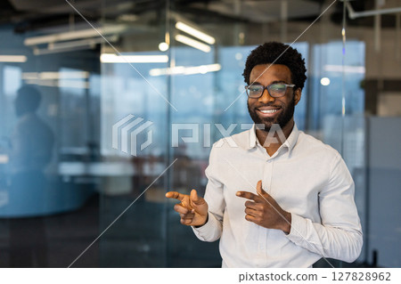 A friendly, smiling man wearing glasses points towards the side in a modern office setting, showcasing his positivity. 127828962