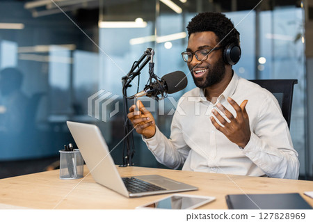A smiling African-American man with glasses records a podcast in a modern office setting. He is using a microphone, headphones, and a laptop. A smiling African-American man with glasses records a podcast in a modern office setting. He is using a microphone, headphones, and a laptop. 127828969