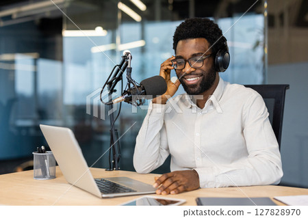 Smiling African American man wearing headphones, sitting at a desk with a microphone and laptop during a podcast recording. Smiling African American man wearing headphones, sitting at a desk with a microphone and laptop during a podcast recording. 127828970