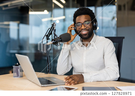 A smiling, handsome man in a podcast setting, wearing glasses and headphones, looking at the camera. There is a microphone, a laptop, and a desk. A smiling, handsome man in a podcast setting, wearing glasses and headphones, looking at the camera. There is a microphone, a laptop, and a desk. 127828971