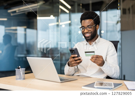 Smiling man wearing glasses is shopping online using his credit card and phone at his desk in his office. Smiling man wearing glasses is shopping online using his credit card and phone at his desk in his office. 127829005