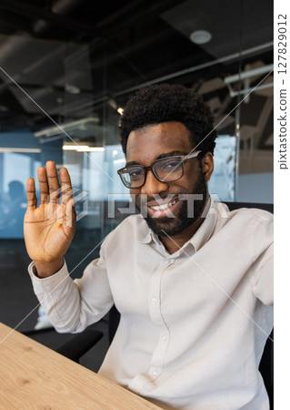 Vertical shot, man waving at smartphone camera, using app for online video call communication. Young businessman inside office at workplace. Vertical shot, man waving at smartphone camera, using app for online video call communication. Young businessman inside office at workplace. 127829012