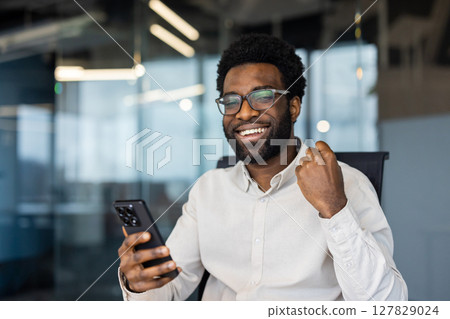 Enthusiastic businessman celebrates success while looking at his mobile phone in a modern office environment, expressing joy and achievement. Enthusiastic businessman celebrates success while looking at his mobile phone in a modern office environment, expressing joy and achievement. 127829024