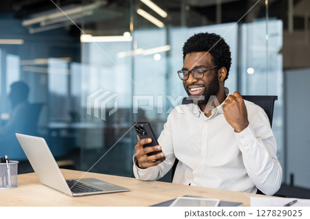 Successful businessman celebrating victory and triumphant achievement results. Man with phone in hands sitting at desk with laptop inside office Successful businessman celebrating victory and triumphant achievement results. Man with phone in hands sitting at desk with laptop inside office 127829025