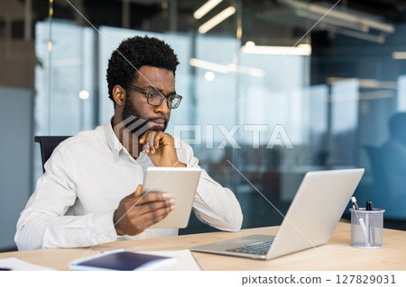 Male thinking businessman working with laptop and tablet computer inside office, man in business shirt viewing online financial report, reading from monitor screen. 127829031
