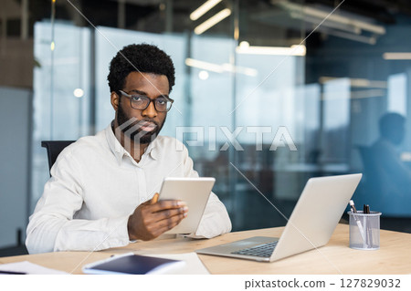 An African American man wearing glasses works at his office desk with a tablet and laptop. He is looking at the camera. An African American man wearing glasses works at his office desk with a tablet and laptop. He is looking at the camera. 127829032