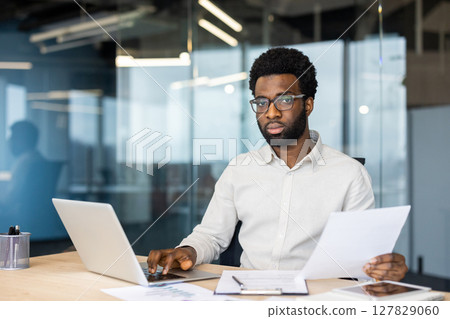 A focused professional balances reviewing documents and working on his laptop in a modern office setting, showcasing productivity and diligence. A focused professional balances reviewing documents and working on his laptop in a modern office setting, showcasing productivity and diligence. 127829060